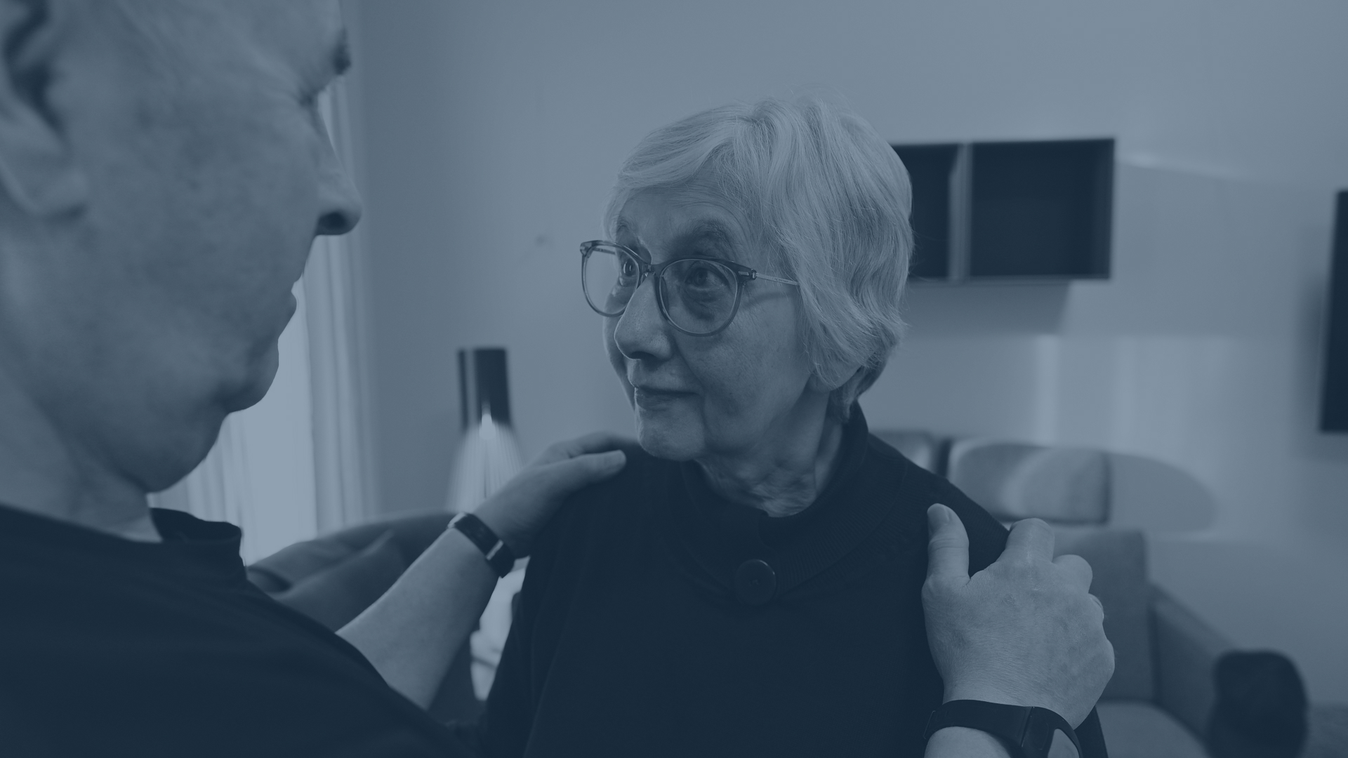An older woman with glasses looks at someone whose hands rest on her shoulders